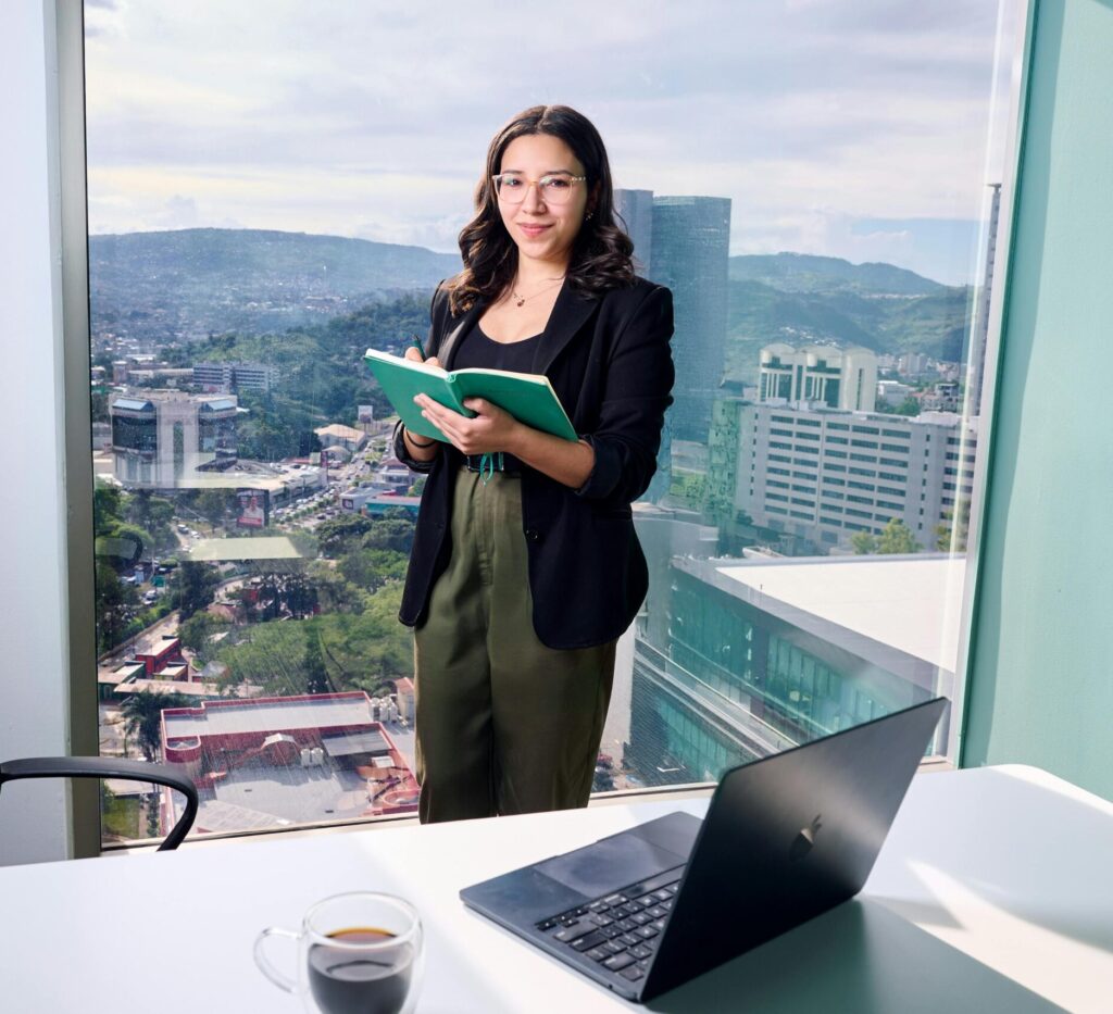 Businesswoman in office holding notebook by window overlooking cityscape.